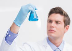 Ein junger Wissenschaftler mit weißem Kittel und Handschuhen hält ein Becherglas mit einer blauen Flüssigkeit in die Höhe