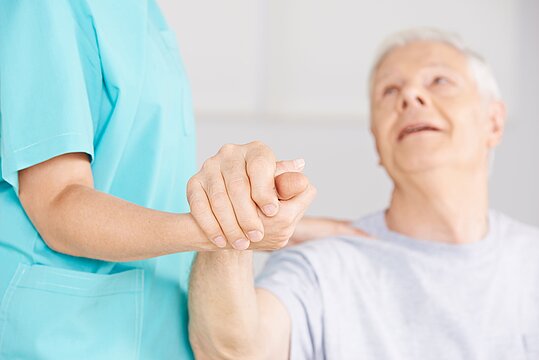 Foto: Eine weibliche Pflegekraft hält die Hand eines sitzenden Mannes.