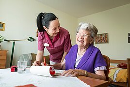 Foto: Eine Pflegerin wendet sich einer Seniorin zu, die an einem Wohnzimmertisch sitzt und Kreuzworträtsel löst