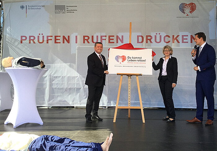 Bundesgesundheitsminister Hermann Gröhe und Dr. Heidrun Thiass, Leiterin der Bundeszentrale für gesundheitliche Aufklärung, stehen auf der Bühne vor einer Staffelei mit einem großen Logo.