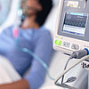 Beatmungsmonitor im Vordergrund, dahinter liegt eine Patientin im Krankenhausbett mit Sauerstoffbeatmungsgerät