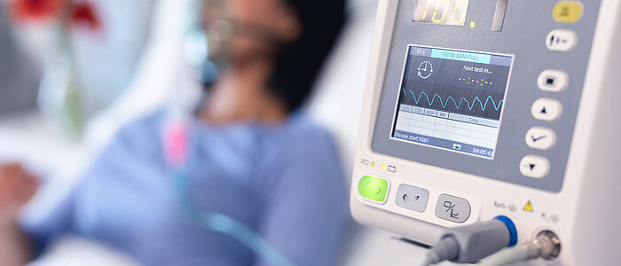 Beatmungsmonitor im Vordergrund, dahinter liegt eine Patientin im Krankenhausbett mit Sauerstoffbeatmungsgerät