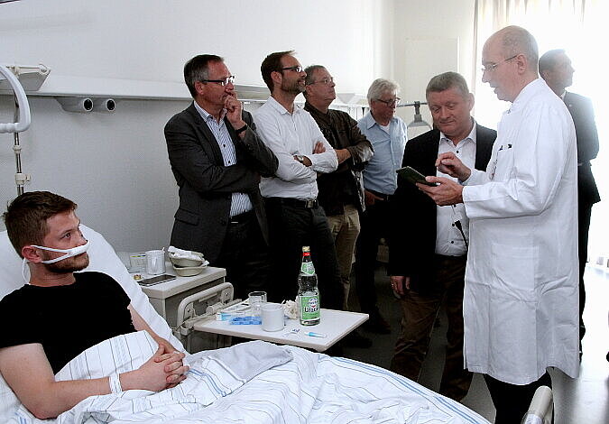 Foto: Bundesgesundheitsminister Gröhe steht neben einem Krankenbett, Prof. Dr. Neumann in weißem Arztkittel hält ihm ein Display entgegen und erklärt etwas, im Hintergrund stehen weitere Personen
