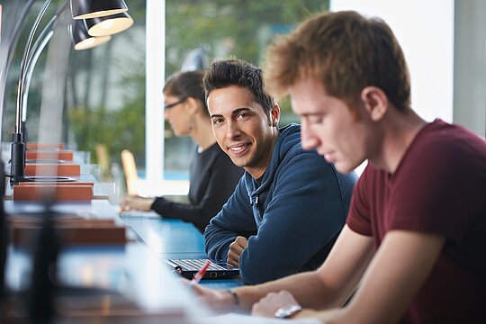 Foto: Drei junge Personen, teilweise schreibend, an Arbeitsplatz mit Leselampe, Laptop und Notizblock in Bibliothek oder ähnlichem.