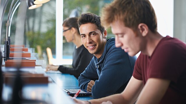 Foto: Drei junge Personen, teilweise schreibend, an Arbeitsplatz mit Leselampe, Laptop und Notizblock in Bibliothek oder ähnlichem.