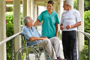 Eine Pflegerin in grünem Kittel steht mit zwei älteren Herren, einer davon im Rollstuhg in einem Gang außerhalb eines Gebäudes.