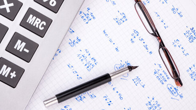 Foto: Taschenrechner, Brille, Stift sowie Berechnungen auf kariertem Notizblock.