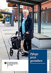 Eine alte Frau steht mit ihrem Rollator an einer Bushaltestelle.