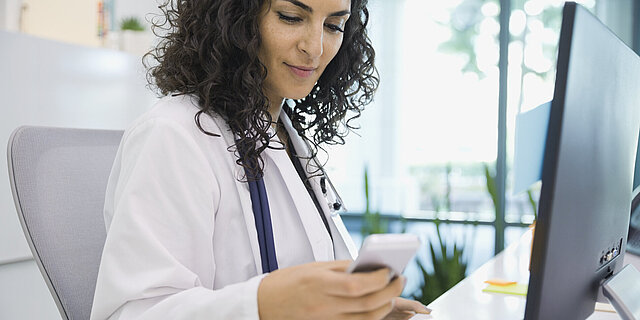 Eine Ärztin sitzt an ihrem Schreibtisch mit Smartphone in der Hand und Stetoskop um ihren Hals
