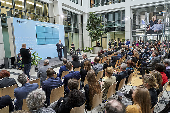 Foto: Im Vordergrund Stulreihen mit Zuschauern, im Hintergrund die Bühne, auf der Jens Spahn spricht, am Seitenrand ein Fotograf.