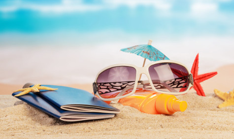 Sommerzeit ist Reisezeit (Bildquelle: Shutterstock/Laboko) Symbolfoto: Eine Zusammenstellung von Sommergegenständen wie einer Sonnenbrille, einer Sonnenmilchflasche, einem Papierschirmchen, mehreren Seesternen und zwei kleinen blauen Heften auf sandigem Grund vor blauem Himmel