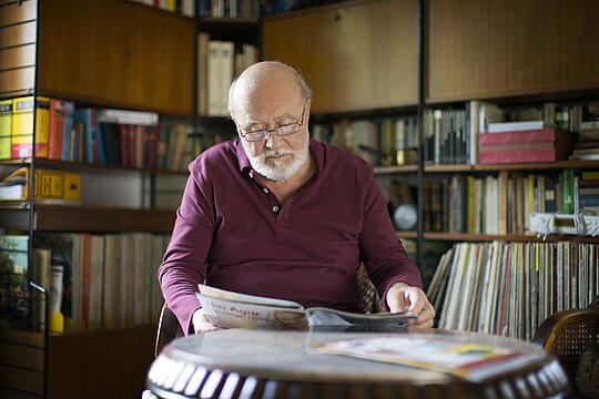 Foto: Mann mit Brille, Halbglatze und grauem Kurzbart in Wohnzimmer oder Bibliothek eine Broschüre oder Zeitschrift lesend