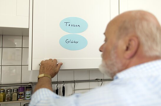 Foto: Ein alter Mann öffnet eine weiße Küchenschranktür auf denen zwei Klebezettel mit den Aufschriften Tassen und Gläser zu sehen sind.