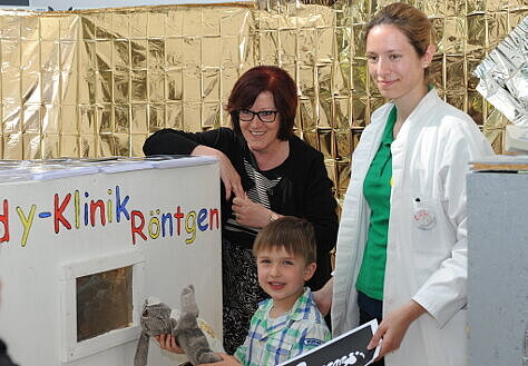Foto: Die Parlamentarische Staatssekretärin Ingrid Fischbach steht mit einem Jungen und dessen Kuschelhasen sowie einer Ärztin an einem Spielzeug-Röntgen-Gerät.