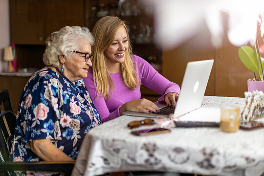 Eine pflegende Angehörige sitzt mit einer älteren Dame vor einem Laptop.