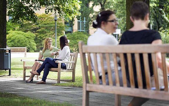 Vier Personen sitzen paarweise auf Bänken im Park und unterhalten sich, im Hintergrund ein Gebäude.