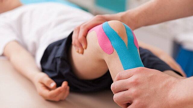 Ein Physiotherapeut führt bei einem Kind eine Knie-Übung durch. Um das Knie sind zwei Pflaster geklebt.