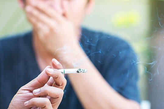 Foto: Eine Zigarette in der Hand einer Person, im Hintergrund eine weitere Person, die sich mit einer Hand Mund und Nase zuhält.