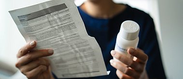 Nahaufnahme einer Frau, die eine Medikamentenflasche in der Hand hält und die Gebrauchsanweisung für die verschriebenen Tabletten aufmerksam liest.