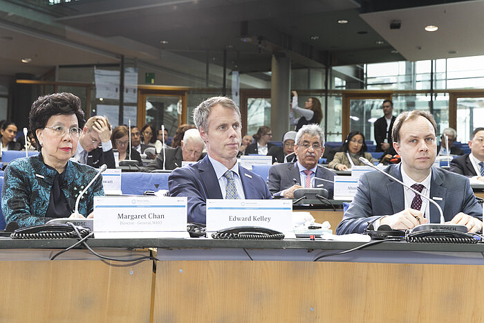 Foto: Margaret Chan, Edward Kelley und Arelijus Veryga sitzen nebeneinander im Veranstaltungssaal, im Hintergrund weitere internationale Gesundheitsminister
