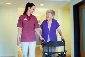 Eine Altenpflegerin läuft mit einer Bewohnerin, die einen Rollstuhl schiebt, über den Flur