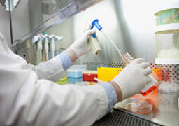 Foto: Jemand in weißem Laborkittel und Handschuhen hantiert mit einem geöffneten Gefäß über einem Labortisch