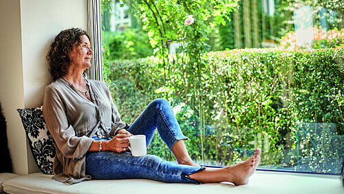 Eine Frau sitzt mit Kaffeetasse und angewinkeltem Bein an einem Panoramafenster und schaut hinaus