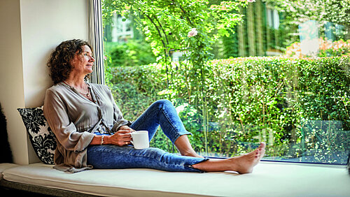 Eine Frau sitzt mit Kaffeetasse und angewinkeltem Bein an einem Panoramafenster und schaut hinaus