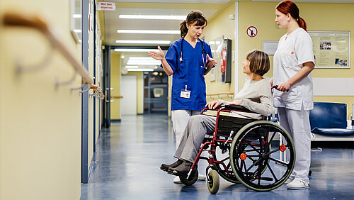 Ärztin erklärt im Krankenhausflur etwas einer älteren Patientin, die im Rollstuhl sitzt