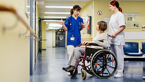 Ärztin erklärt im Krankenhausflur etwas einer älteren Patientin, die im Rollstuhl sitzt