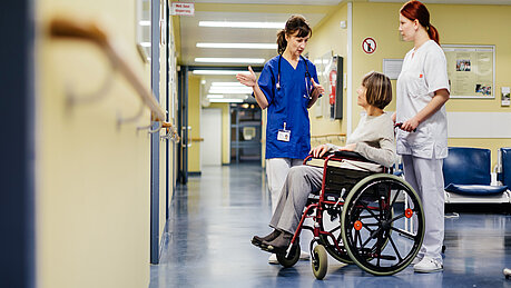 Ärztin erklärt im Krankenhausflur etwas einer älteren Patientin, die im Rollstuhl sitzt