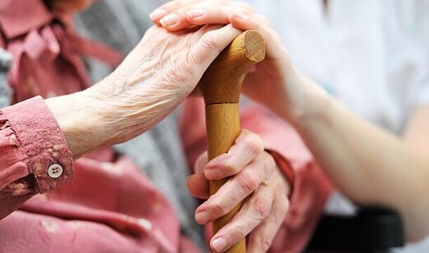 Foto: Hände einer älteren Frau umfassen das Ende des Gehstocks, die Hand einer Betreuungskraft liegt obendrauf