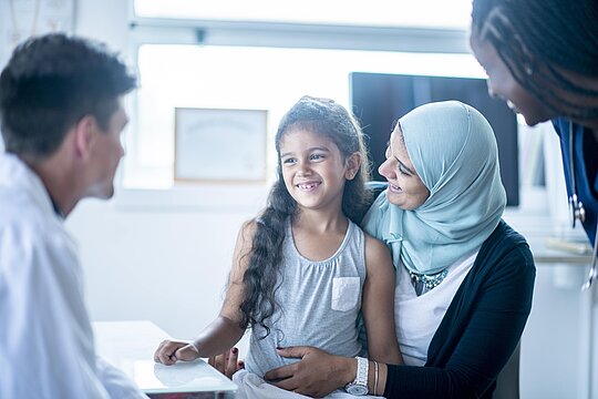 Foto: lächelndes Mädchen mit kopftuchtragender Mutter sowie nebenstehender Frau mit Rasterzöpfen und dunkler Hautfarbe im Gespräch mit einem Arzt mit heller Hautfarbe