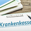 Foto: diverse Berichte mit Spiralbindung auf einem Tisch sowie Klemmbrett, Kugelschreiber, großer Schriftzug "Krankenkasse".