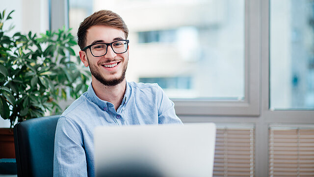Ein Mann sitzt mit Laptop im Homeoffice und lächelt in die Kamera