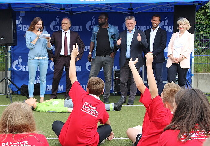 Foto: Minister Gröhe steht auf der Bühne und zeigt den Kindern, die davor sitzen, den "Daumen hoch", neben dem Minister stehen unter anderem Gerald Asamoah und Dr. Heidrun Thaiss