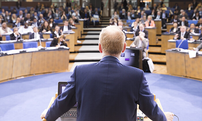 Foto: Sir Liam Donaldson steht mit dem Rücken zum Betrachter an einem Rednerpult, im Bildhintergrund sind die zuhörenden Kongressteilnehmer zu sehen