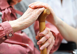 Foto: Eine ältere Dame hält einen Gehstock, auf ihrer Hand liegt die Hand einer jüngeren Frau in weißem Kittel