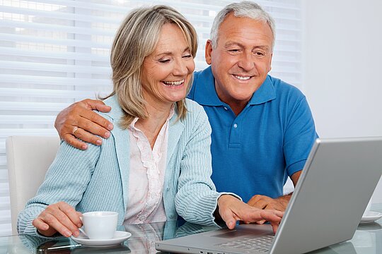 Foto: Mann mit Frau schauen lächeln auf ein Tablet. Die Frau berührt mit den Fingern der einen Hand den Bildschirm und hält mit der anderen Hand eine Tasse.