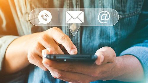 EIne Person hält ein Handy ind en Händen und über dem Handy schweben drei Symbole (eine Sprechblase, ein Briefcurvert und ein at)