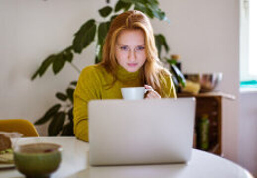 Foto: Frau mit Tasse in der Hand am Tisch sitzend, blickt in den Laptop. 