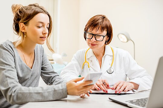Foto: Ärztin und Patienin im Gespräch, schauen auf das Handy der Patientin
