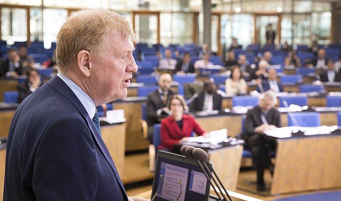 Foto: Sir Liam Donaldson ist seitlich fotografiert am Rednerpult zu sehen, im Hintergrund hören ihm die Kongressteilnehmer zu