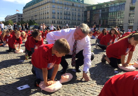 Bundesgesundheitsminister Hermann Gröhe kniet im Freien neben einem Jungen. Beide üben Maßnahmen zur Wiederbelebung an einer Puppe.