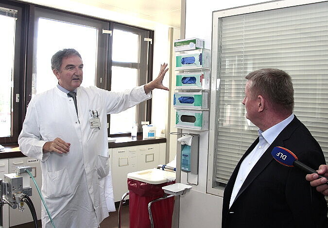 Foto: Minister Gröhe in einem Krankenhausraum, in dem ihm ein Arzt etwas erklärt und auf Boxen mit Einmalhandschuhen zeigt