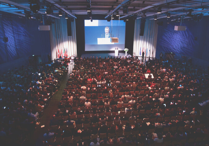 Foto: Ein gefüllter, abgedunkelter Sitzungssaal, Hermann Gröhe steht am Rednerpult und wird zusätzlich für die Zuschauer auf eine Leinwand projeziert