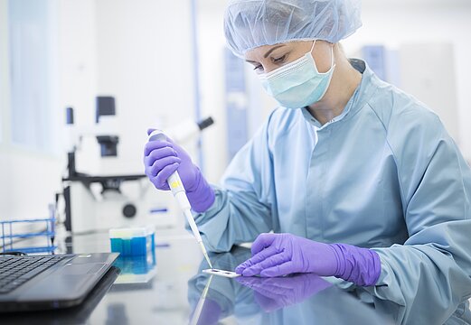 Forscherin im Labor mit Kopfbedeckung, Maske, Handschuhen und Pipette in der Hand