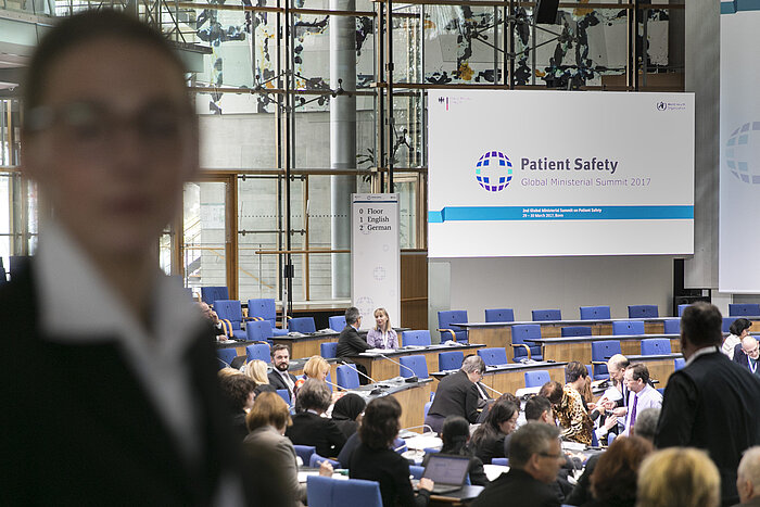 Foto: Blick in den Veranstaltungssaal; im Vordergrund eine Teilnehmerin, im Hintergrund Sitzreihen und eine Präsentationsfolie mit dem Patient Safety Summit Logo