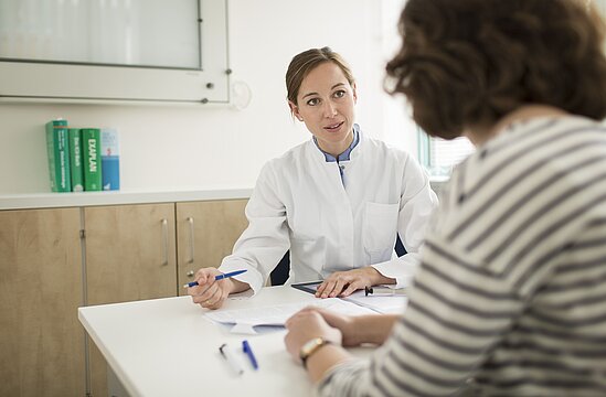 Foto: Ärztin, einen Kugelschreiber in der Hand haltend, am Tisch und im Gespräch mit Patientin.