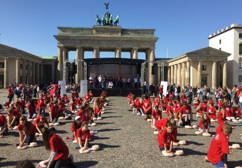 Im Vordergrund üben Kinder in roten T-Shirts Wiederbelebungsmaßnahmen an Puppen. Im Hintergrund ist eine Bühne vor dem Brandenburger Tor zu sehen.